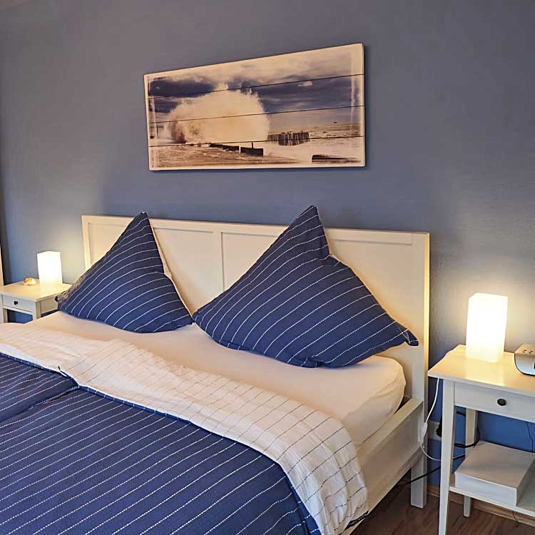 Schlafzimmeransicht: Doppelbett, zwei Nachttische mit eigenen Nachtischlampen. Hinter dem Bett eine blaue Wand und ein Nordseebild auf Holz