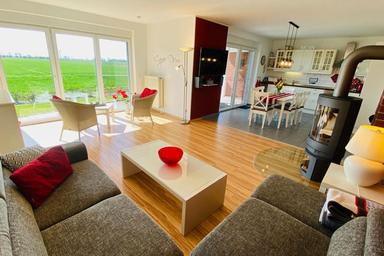 Das lichtdurchflutete Wohnzimmer vom Ecksofa aus fotografiert: man sieht das Ecksofa mit Tisch und gemütlicher Beleuchtung, eine zusätzliche Sitzecke mit 2 Stühlen und Stehlampe, den Kamin, den Essbereich mit Küche